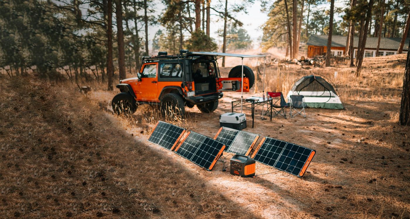 Cuatro paneles solares portátiles cargando una estación de energía junto a un todoterreno y una tienda de campaña